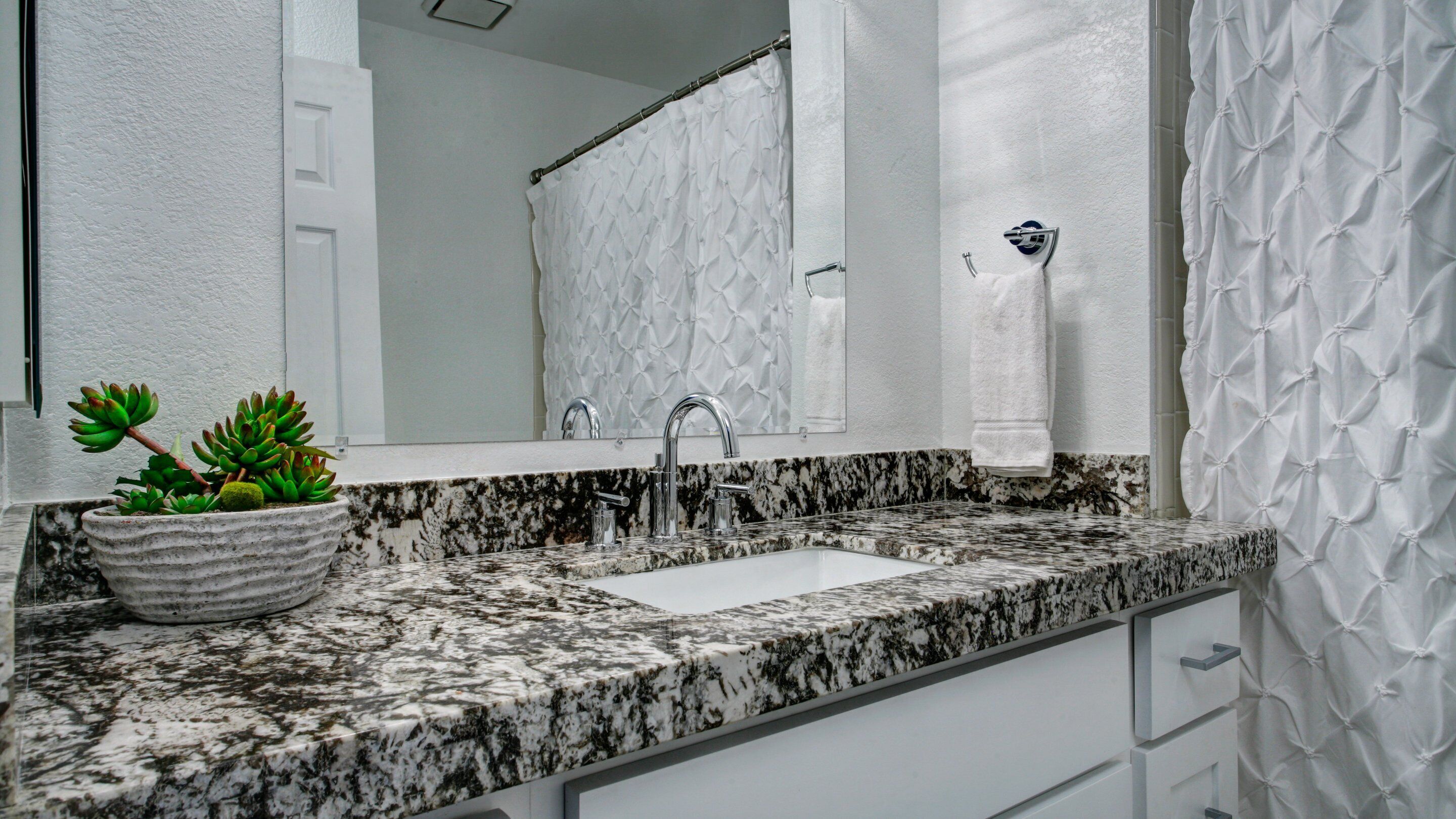 A bathroom with a granite countertop, a white sink, a mirror, a shower curtain, and a potted plant.