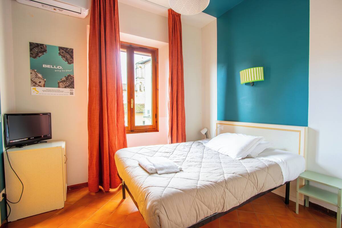 Una habitación de hotel con una cama, un televisor sobre un mueble bajo, una ventana con cortinas y una pared con un cartel.