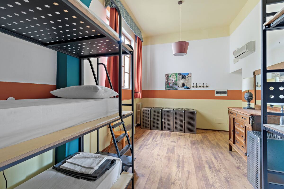 Habitación con literas, suelo de madera, escritorio con cajones, radiador y estante de pared con adornos.