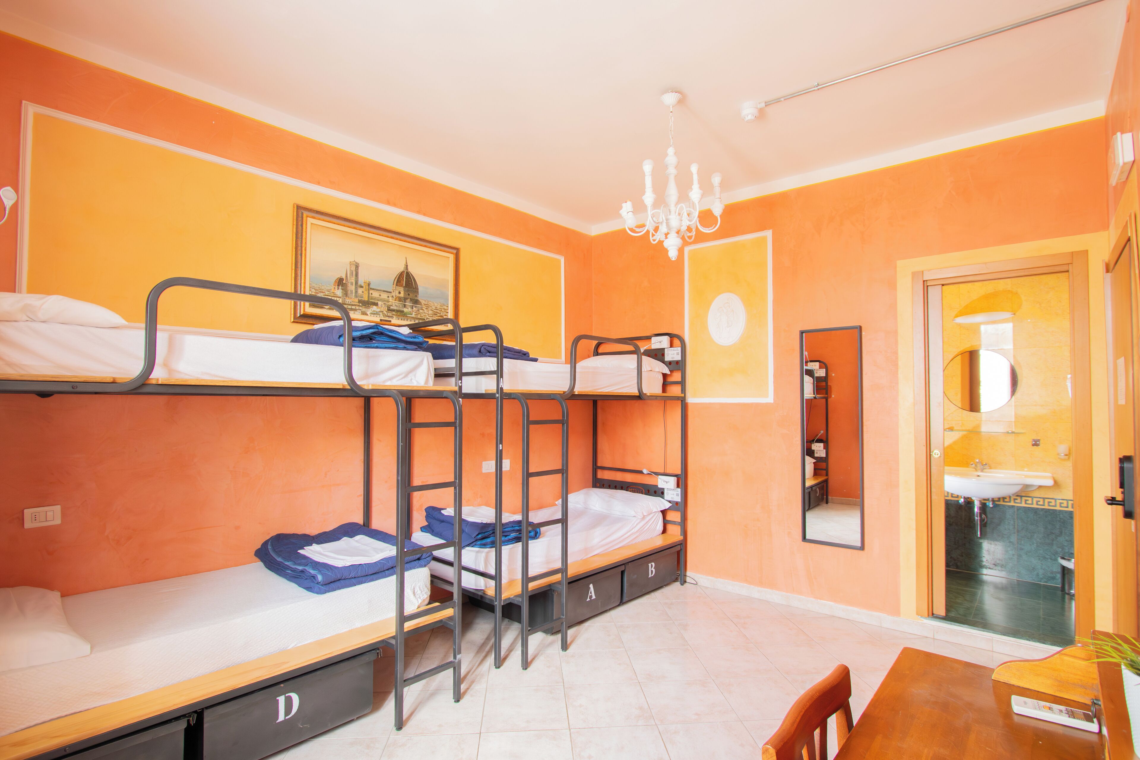 A dormitory room with bunk beds, a chandelier, a painting, and a bathroom visible through an open door.