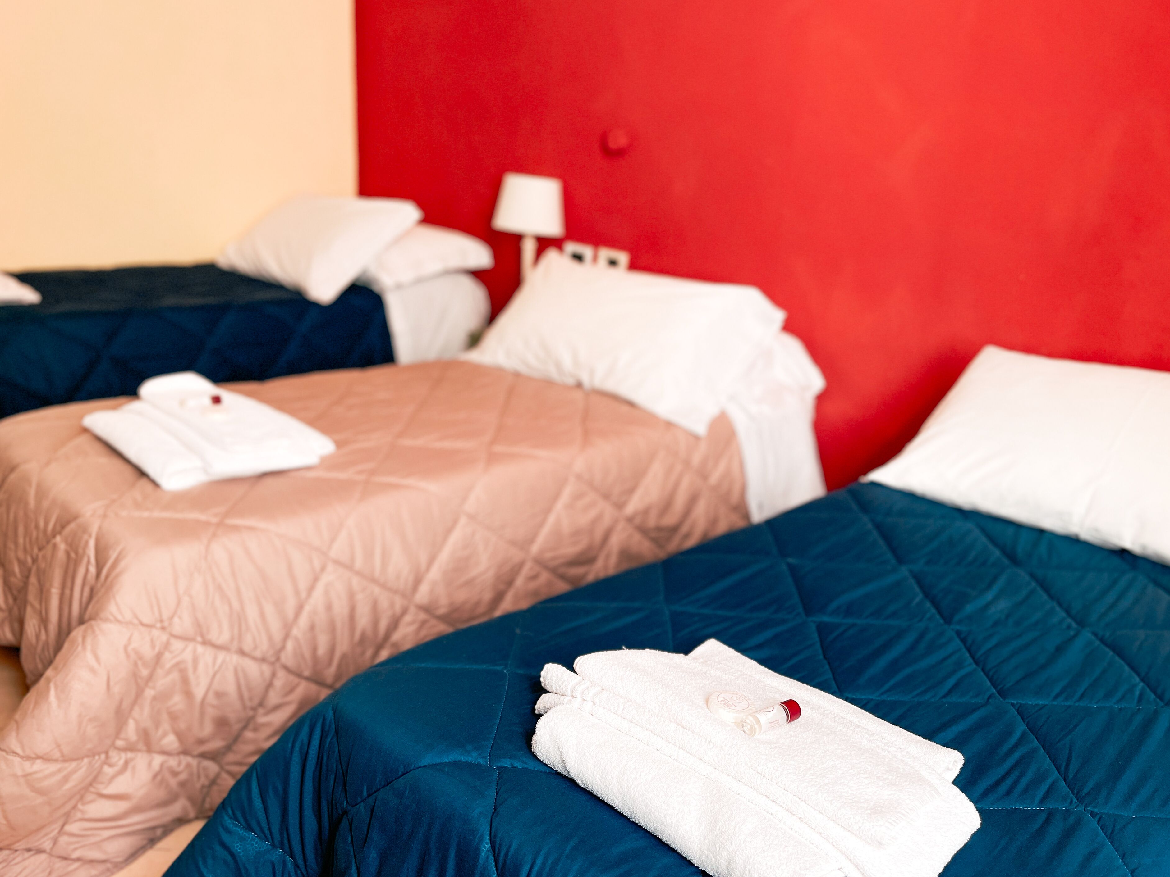 Two beds with white towels folded on them, a red wall, and a small table with a lamp.