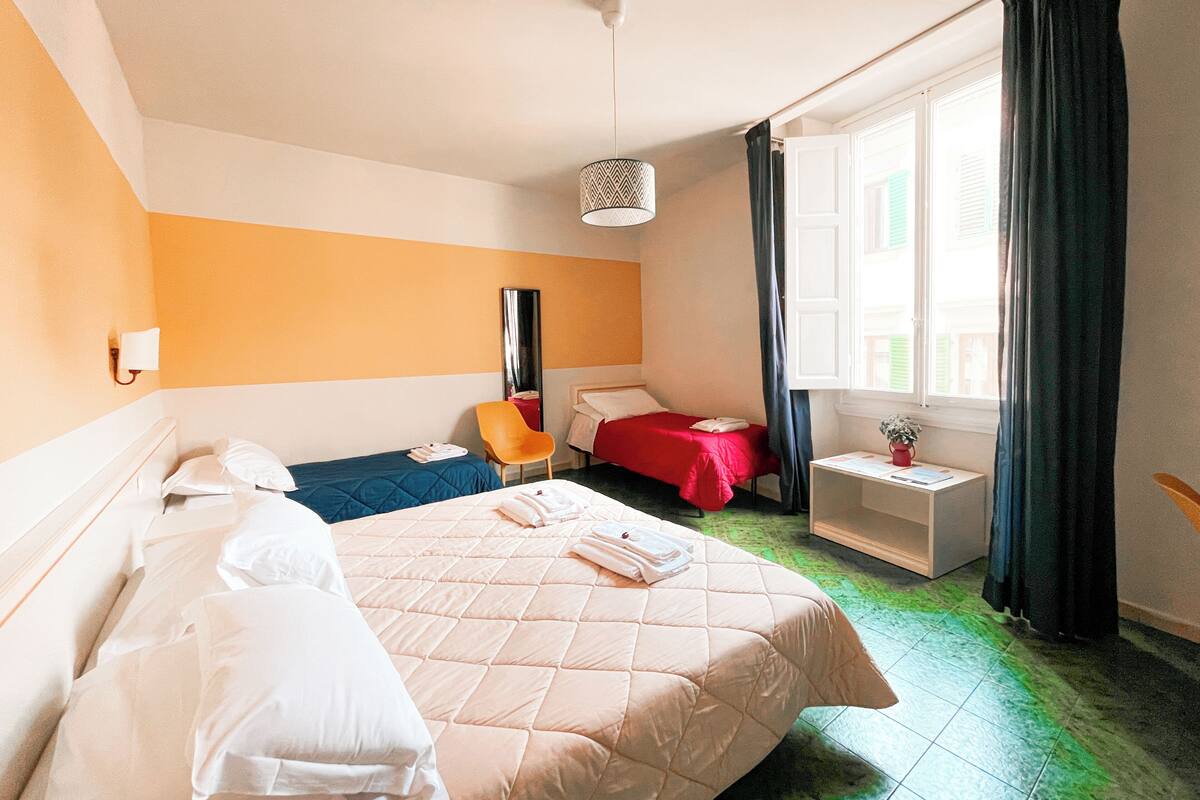 Habitación de hotel con cama, una silla, mesita de noche, espejo y ventana con cortinas.