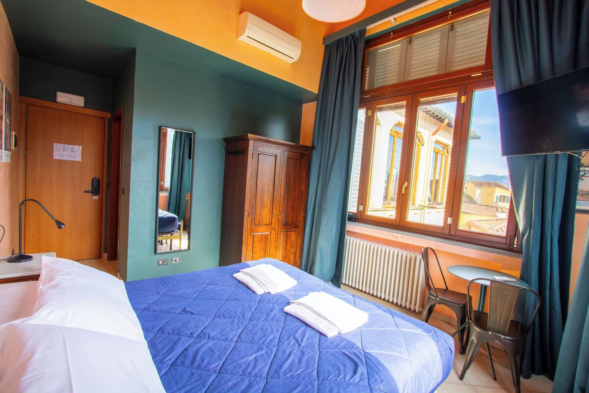 Habitación de hotel con cama, escritorio, silla, espejo, ventana con cortinas y radiador.
