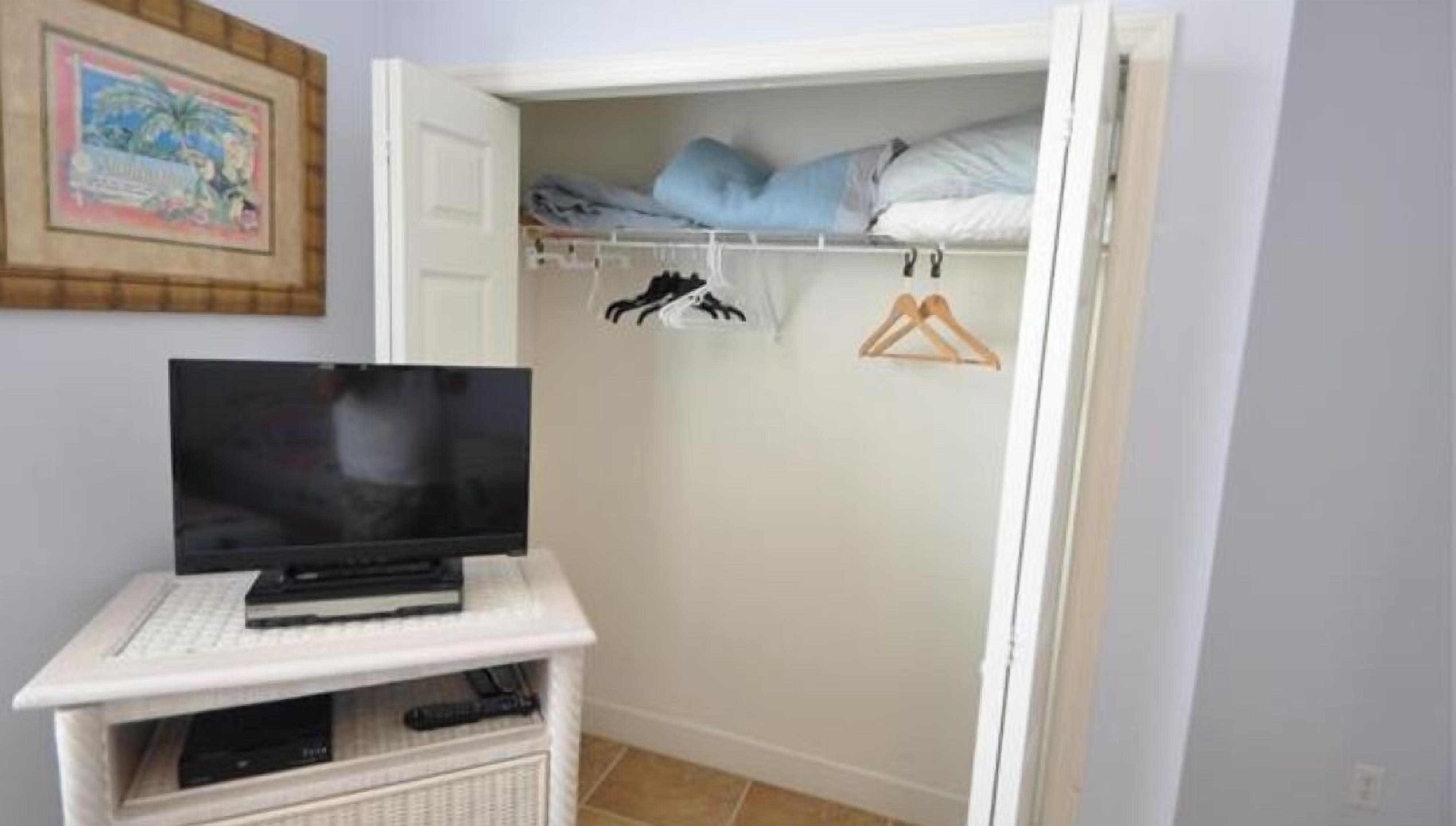 A room with a TV on a white stand, a closet with clothes and hangers, and a framed picture on the wall.