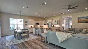 A spacious living area with a grey sofa, a dining table with chairs, and a kitchen with white cabinets and modern appliances.