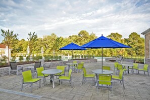 An outdoor patio with round tables and chairs, blue umbrellas, and a pool in the background.
