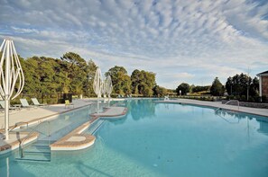 A large outdoor swimming pool with clear blue water, surrounded by lounge chairs and umbrellas, with trees in the background.