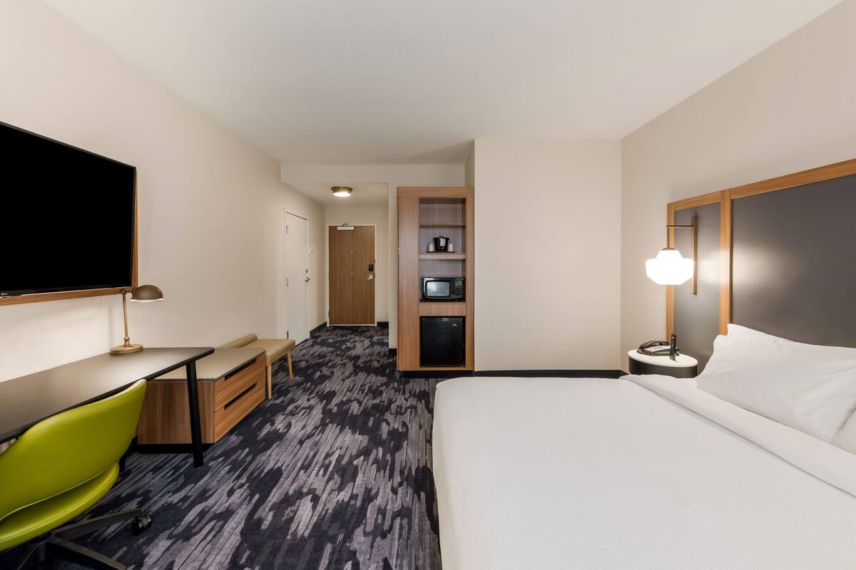 Hotelkamer met een bed, een bureau, een groene stoel, een televisie, een magnetron en een nachtkastje met een lamp.