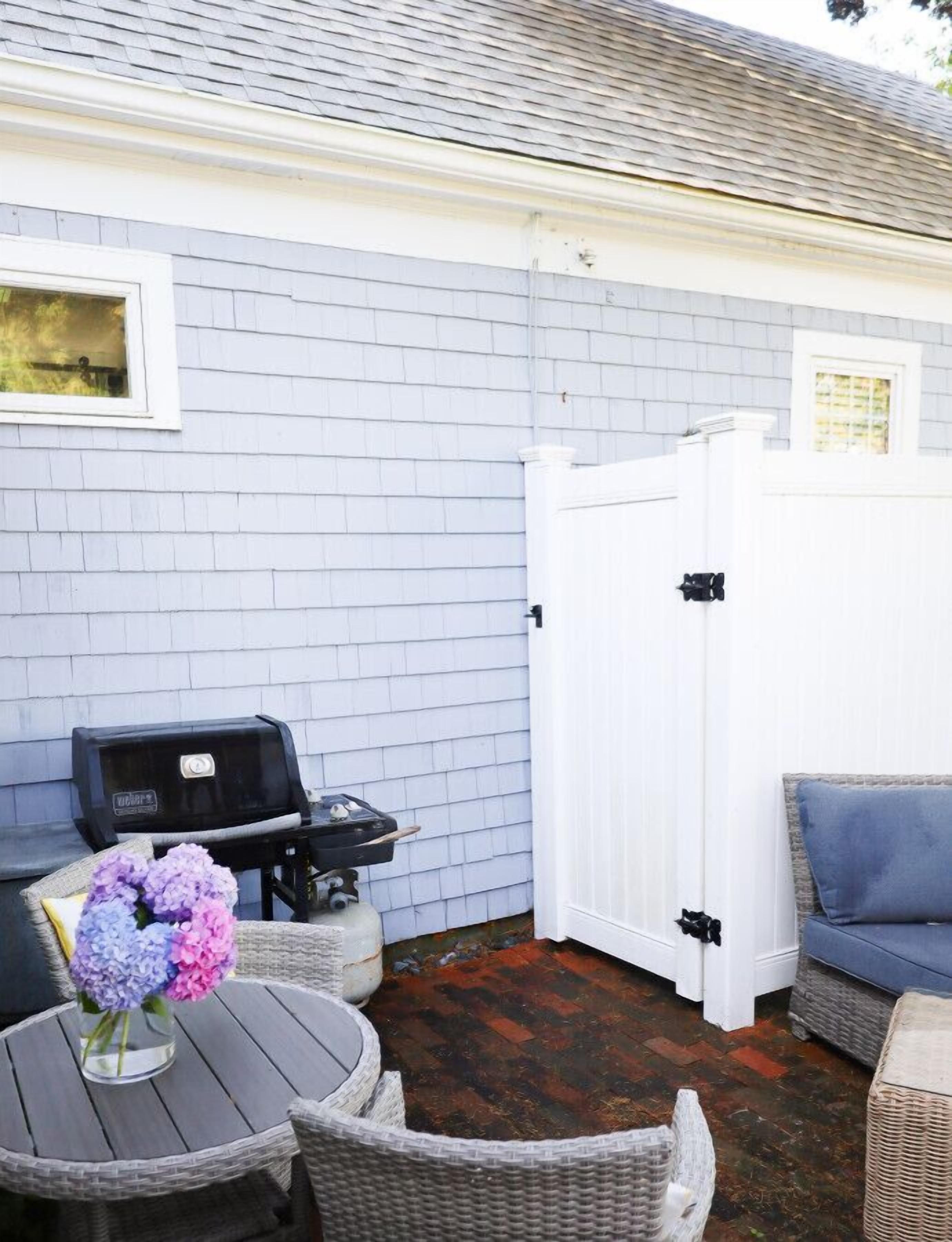 A patio with a grill, a round table, wicker furniture, and a white fence.