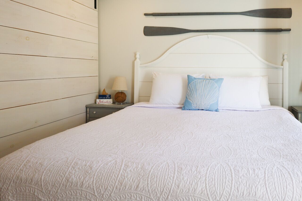 Una cama bien hecha con colcha blanca, cabecera de madera y dos almohadas, una azul y otra blanca.
