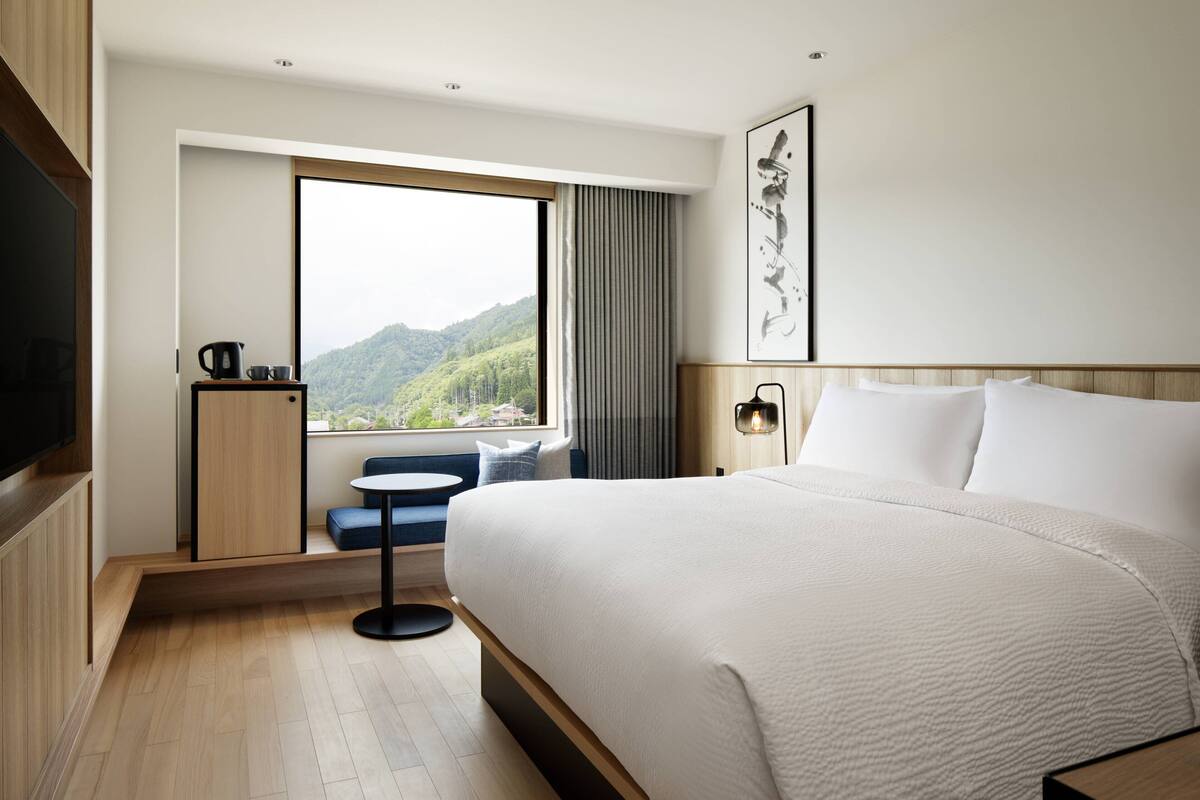 Une chambre d’hôtel moderne avec un grand lit, une petite table avec une bouilloire, une vue sur les montagnes par la fenêtre et un téléviseur à écran plat sur le côté gauche.