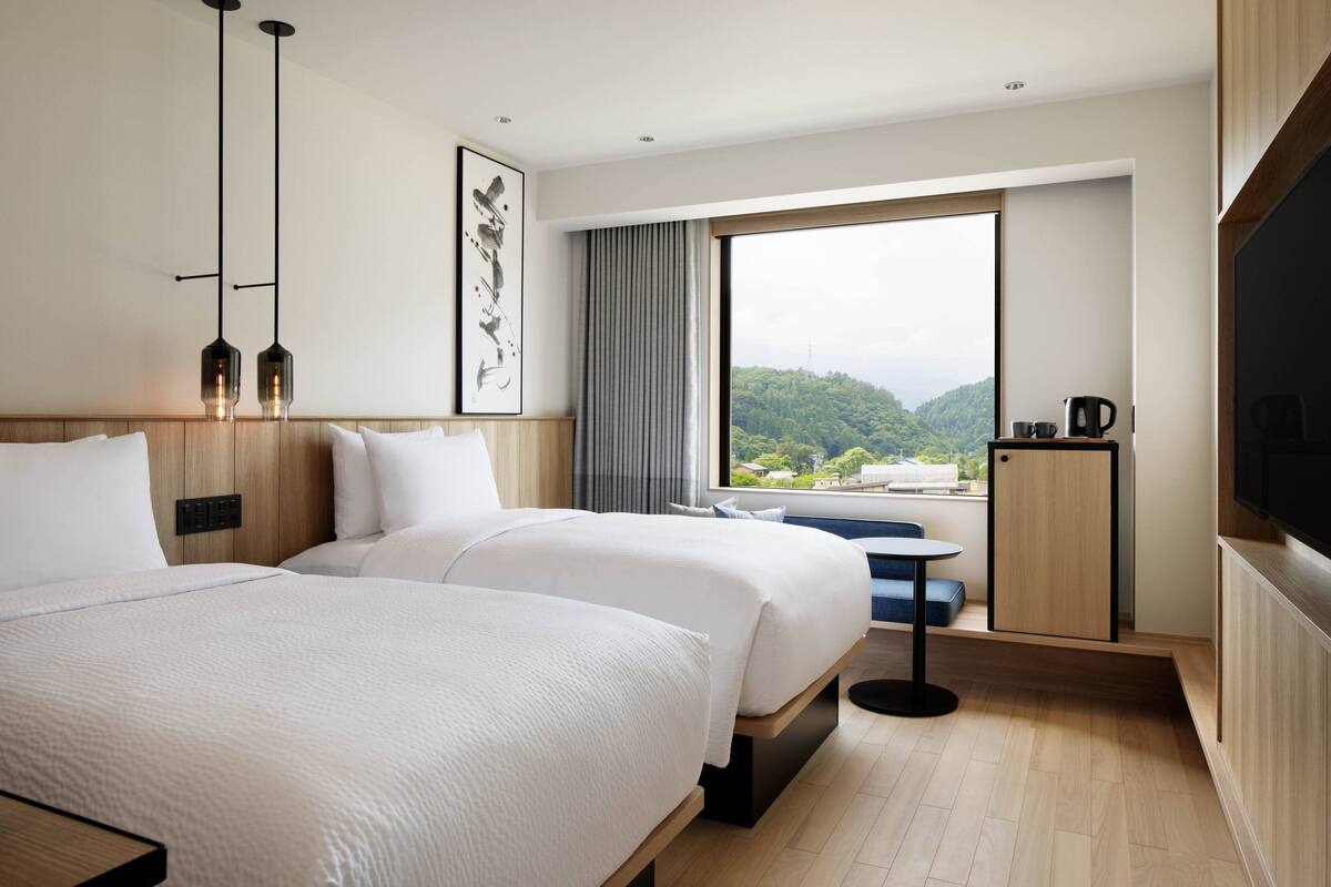 Une chambre d’hôtel avec deux lits, une grande fenêtre donnant sur un espace verdoyant, un téléviseur à écran plat et une petite table avec une machine à café.