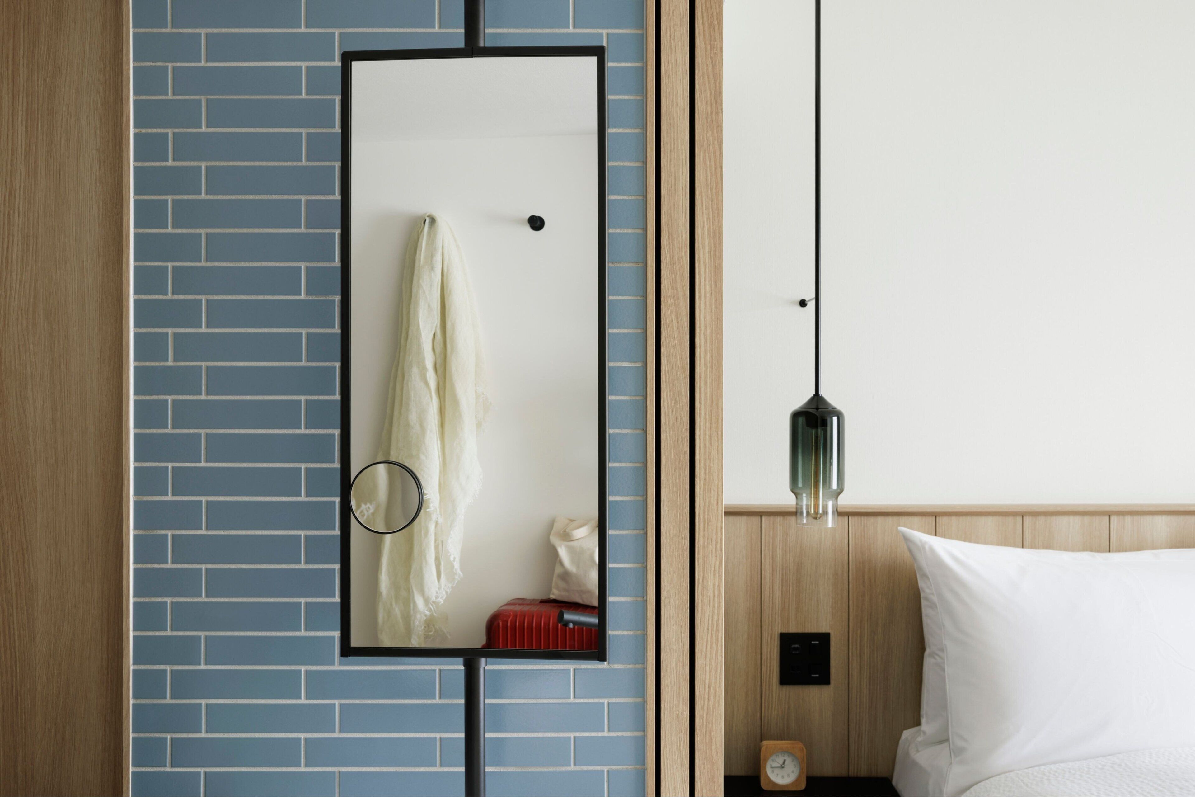 A hotel room with a blue-tiled wall, a wooden headboard, a bed with white bedding, a nightstand with a clock, and a mirror with a towel hanging on it.