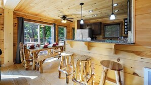 A wooden cabin interior with a dining area, bar stools, a ceiling fan, and a kitchen with modern appliances.