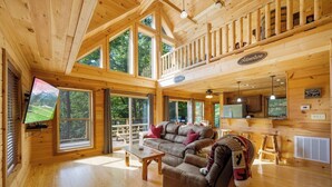 A spacious wooden cabin with a loft bedroom, a cozy living area with a leather sofa, and large windows offering views of the surrounding forest.
