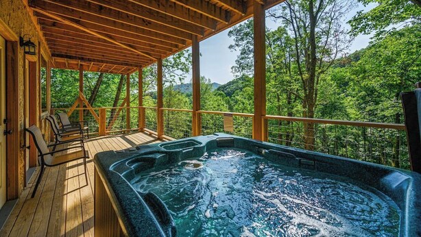 A wooden deck with a hot tub and seating area, surrounded by trees and a mountainous backdrop.