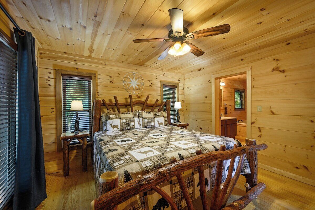 A log cabin bedroom with a bed, a ceiling fan, a compass wall decoration, and a small table with a lamp.