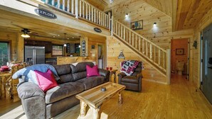 A spacious log cabin interior with a dining area, a living room with a sofa and coffee table, a kitchen, and a staircase leading to an upper level.