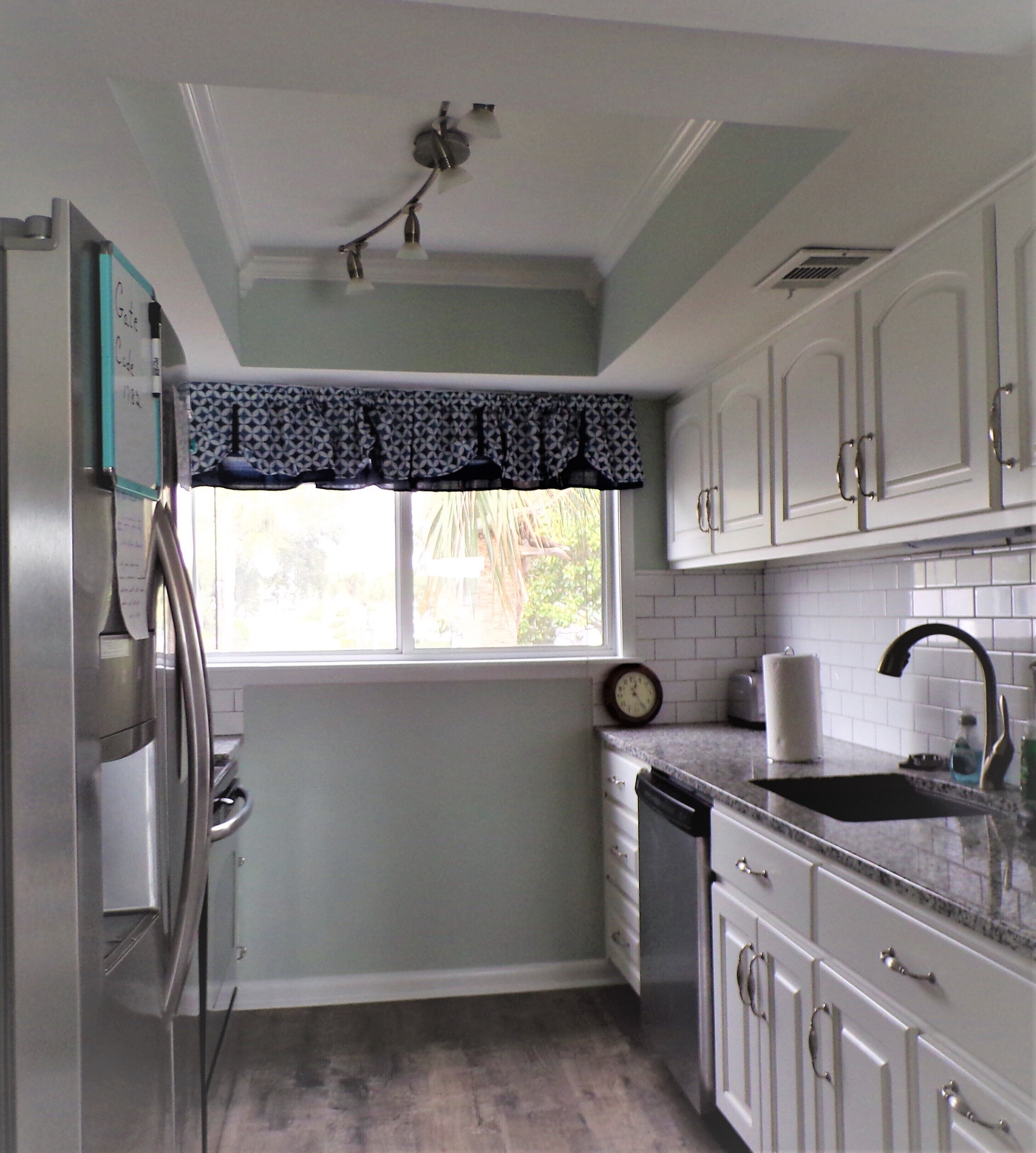Una cocina con armarios blancos, un refrigerador de acero inoxidable, un ventanal con un toldo estampado y un reloj en la pared.