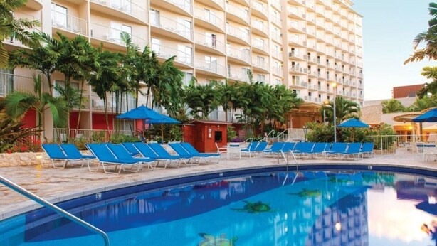 Un espace piscine d’hôtel avec des chaises longues bleues, une piscine à l’eau cristalline et un immeuble à plusieurs étages doté de balcons.