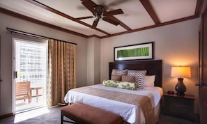 A hotel room with a bed, a ceiling fan, a window with curtains, a framed picture on the wall, and a bedside lamp.