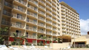Un hôtel à plusieurs étages doté de balcons, d’un espace piscine avec des chaises longues, et un bâtiment rouge en arrière-plan.