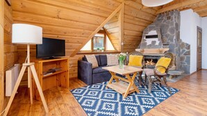Un acogedor interior de cabaña de madera con chimenea de piedra, un sofá, un televisor y una zona de comedor.