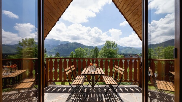 Un balcón con mesa y sillas de madera, un vaso de agua y vistas a montañas y árboles.