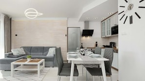 A modern living room with a grey sofa, a dining table set for four, and a kitchen area with a clock on the wall. - Modern Apartment close to the Beach | Balcony | Parking (Darlowo)