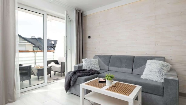 A modern living room with a grey sofa, white coffee table, and a view through a glass door to an outdoor seating area. - Modern Apartment close to the Beach | Balcony | Parking (Darlowo)
