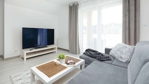 A modern living room with a flat-screen TV on a white stand, a grey sofa, a white coffee table with a plant and a book, and large windows with curtains. - Modern Apartment close to the Beach | Balcony | Parking (Darlowo)