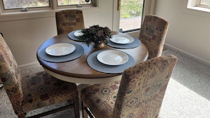 A dining table with four chairs, a vase with greenery, and a view of trees through the window.