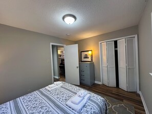 Ein Schlafzimmer mit Bett, Kommode, Schrank mit Klappläden und einem zur Toilette führenden Gang.
