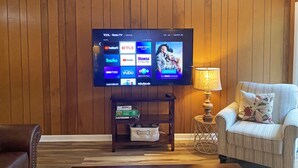 A living room with a flat-screen TV mounted on a wooden wall, a small wooden side table with a lamp, a cushioned armchair, and a coffee table. - Cozy Lake Hartwell Cottage near Tugaloo State Park (Lavonia)