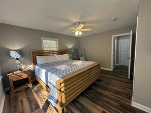 A bedroom with a wooden bed, a ceiling fan, a bedside lamp, a window with blinds, and a door leading to another room.