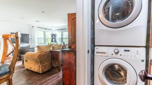 A laundry room with stacked washers and dryers, a wooden chair, a small table, and a sofa in a bright room with large windows.