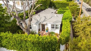A white house with a grey roof, surrounded by lush green trees and hedges.