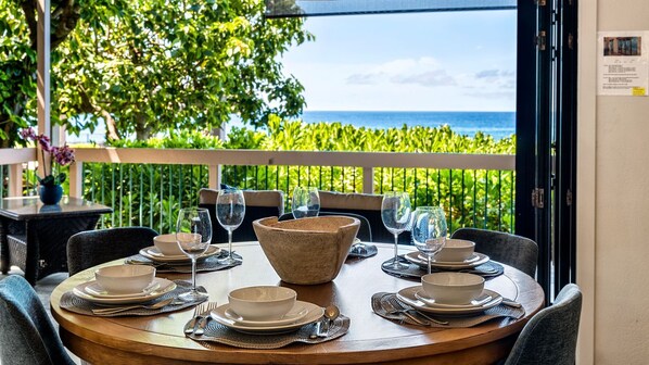 A dining table set for six with a large bowl centerpiece, surrounded by chairs and plates, with a view of the ocean through a glass door. - 🏖️Ocean Front Views | Central AC | 3 Complex Pools (Kailua Kona)