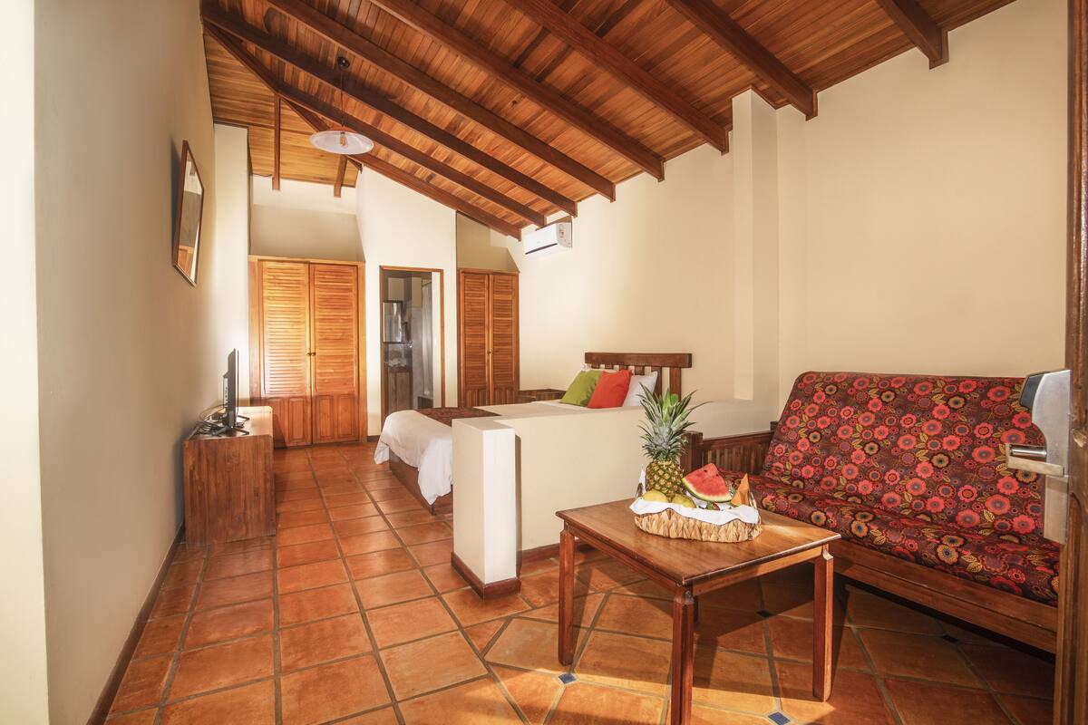 A room with a bed, a sofa, a wooden table with fruits, and a tiled floor.