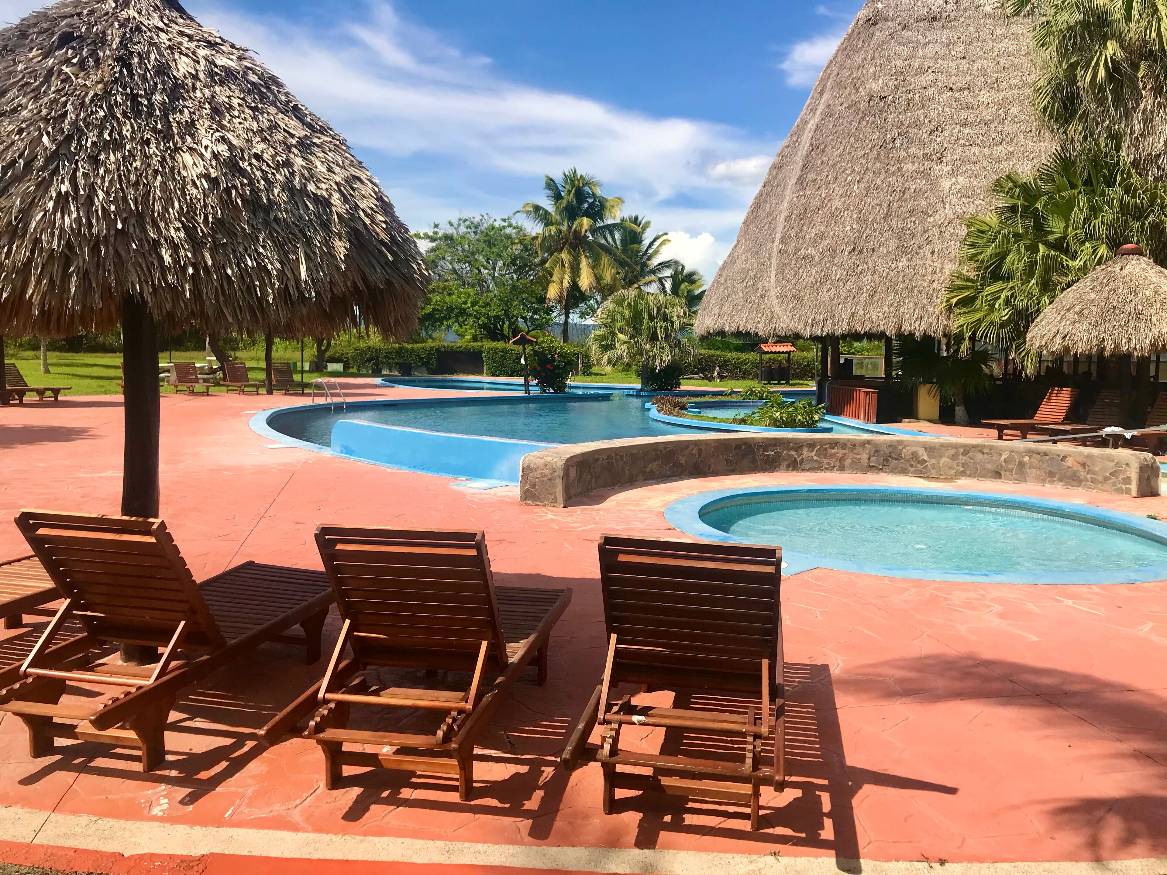 Zona de piscina en un resort con estructuras de techo de paja, sillas de descanso y un cielo azul despejado.