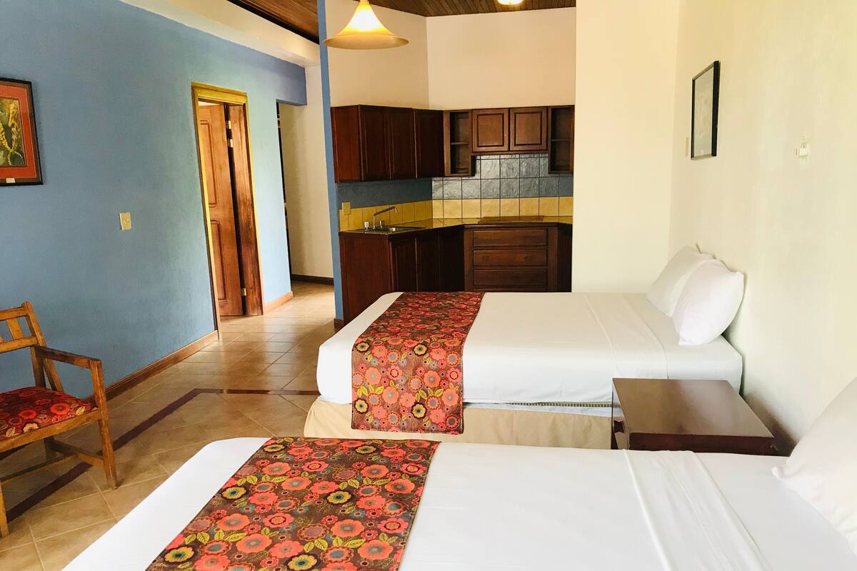 A hotel room with two beds, a small table, a chair, a kitchenette, and a door leading to another room.