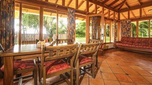 A wooden dining area with a table and chairs, large windows, and a red patterned sofa. - Nandel Beach Resort (La Cruz)