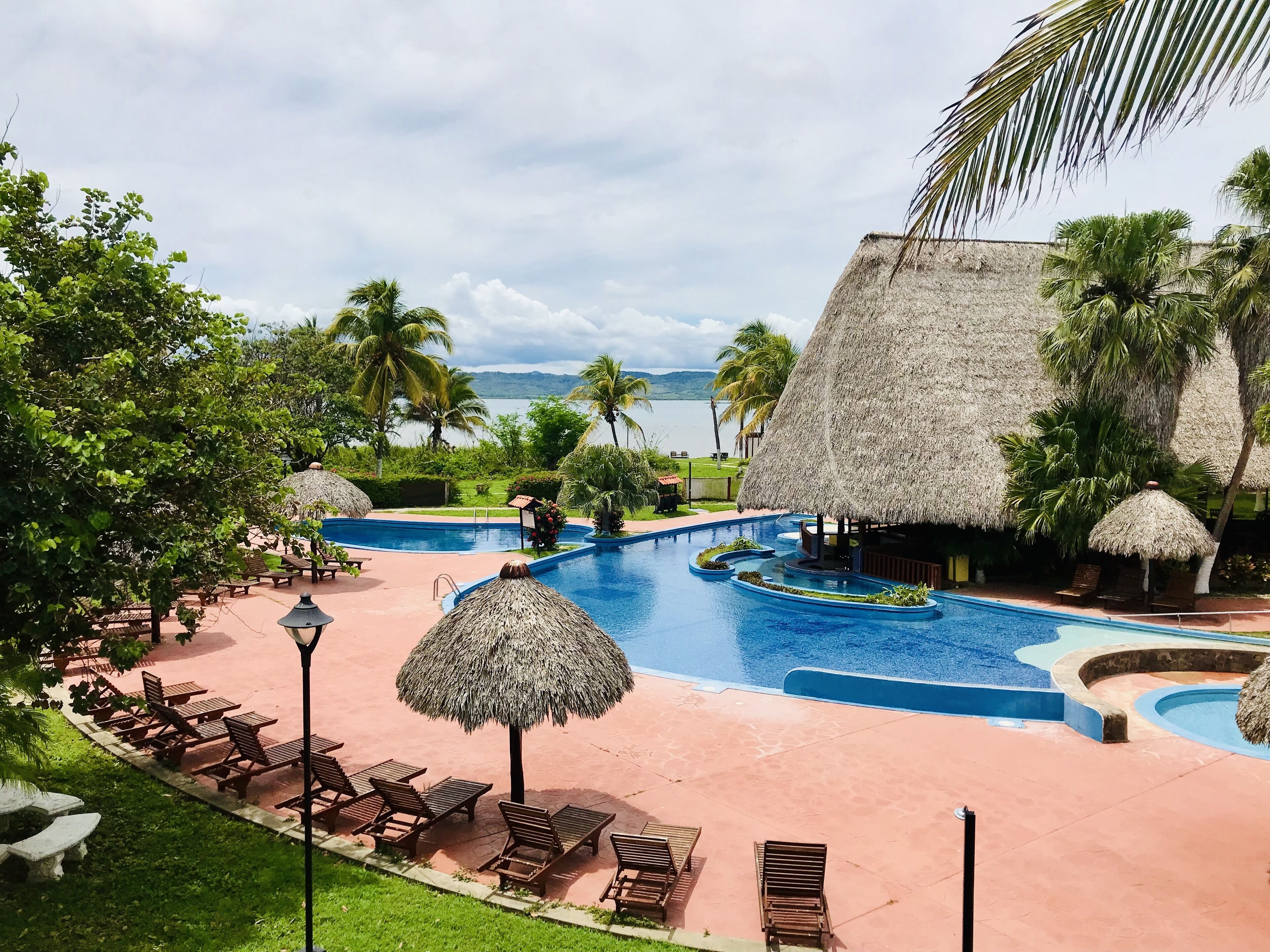 A resort pool area with thatched-roof structures, lounge chairs, and a view of the sea.