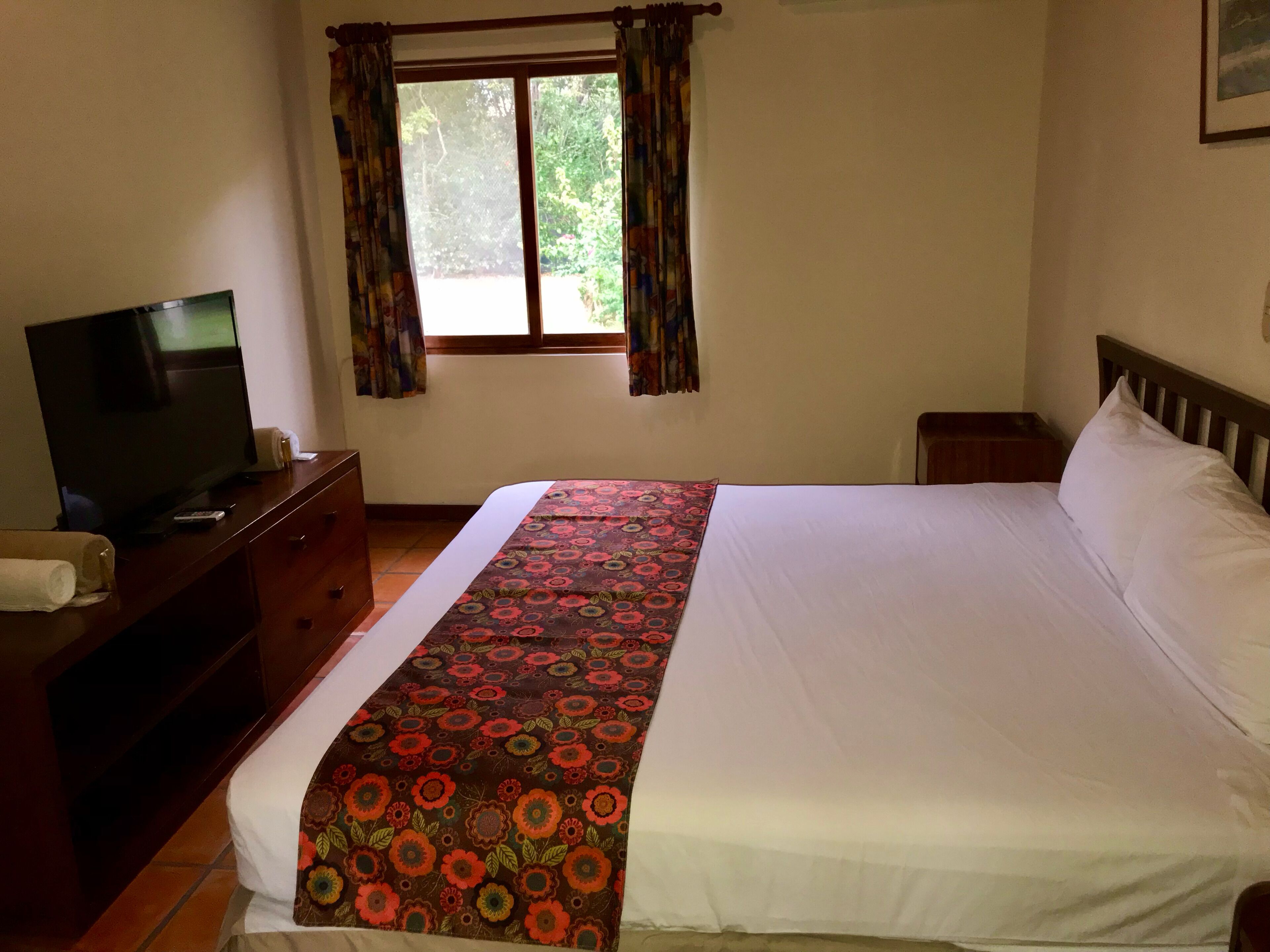 Habitación de hotel con cama, televisor sobre una cómoda de madera, ventana con cortinas y una mesita de noche pequeña.