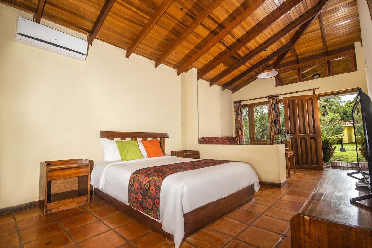 A bedroom with a wooden ceiling, a bed with a floral bedspread, a nightstand, and a window with curtains.