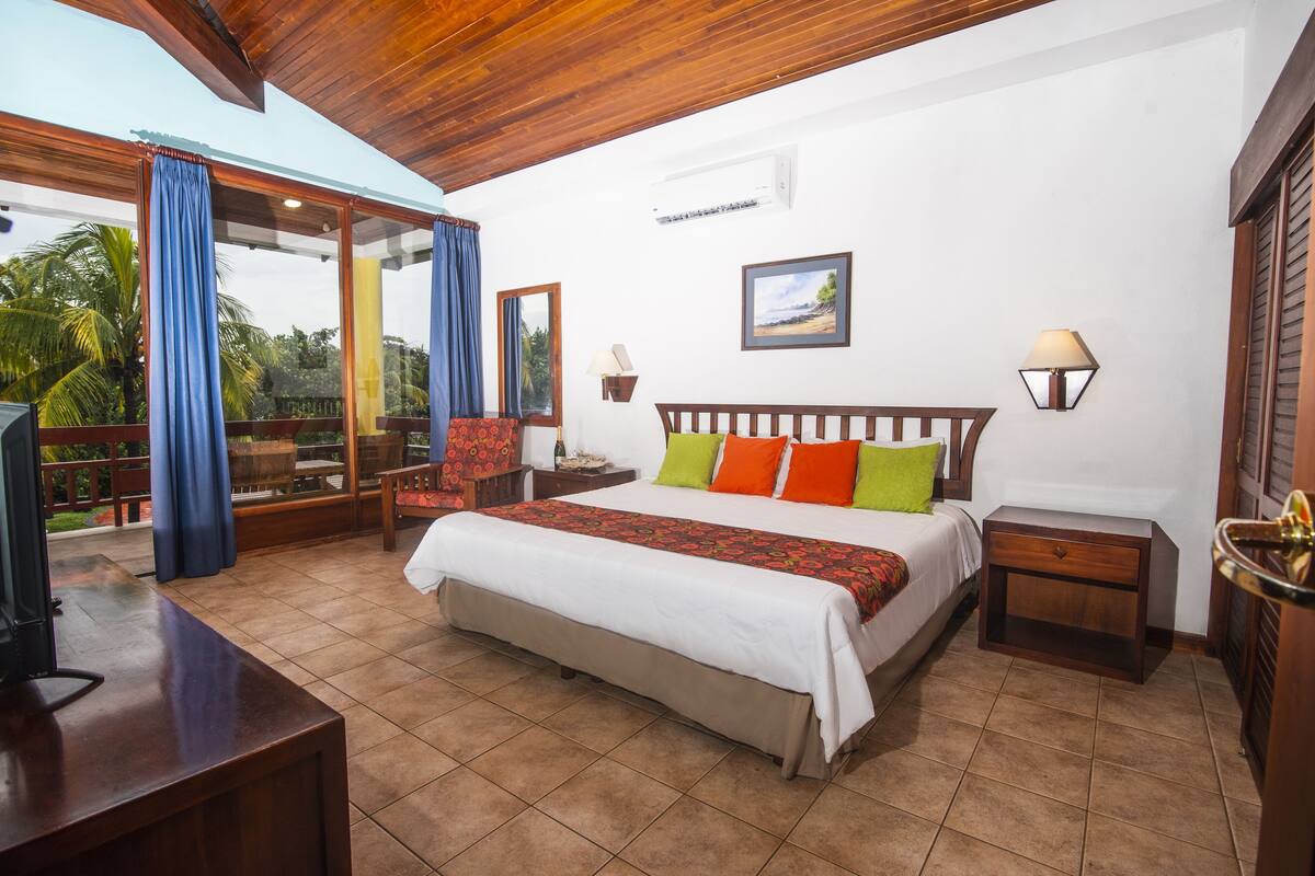 A bedroom with a large bed, a wooden headboard, a nightstand, a desk, and a window with curtains.