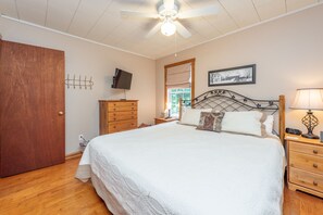 A bedroom with a bed, wooden dresser, nightstand, lamp, ceiling fan, TV, and a window with a view of trees.