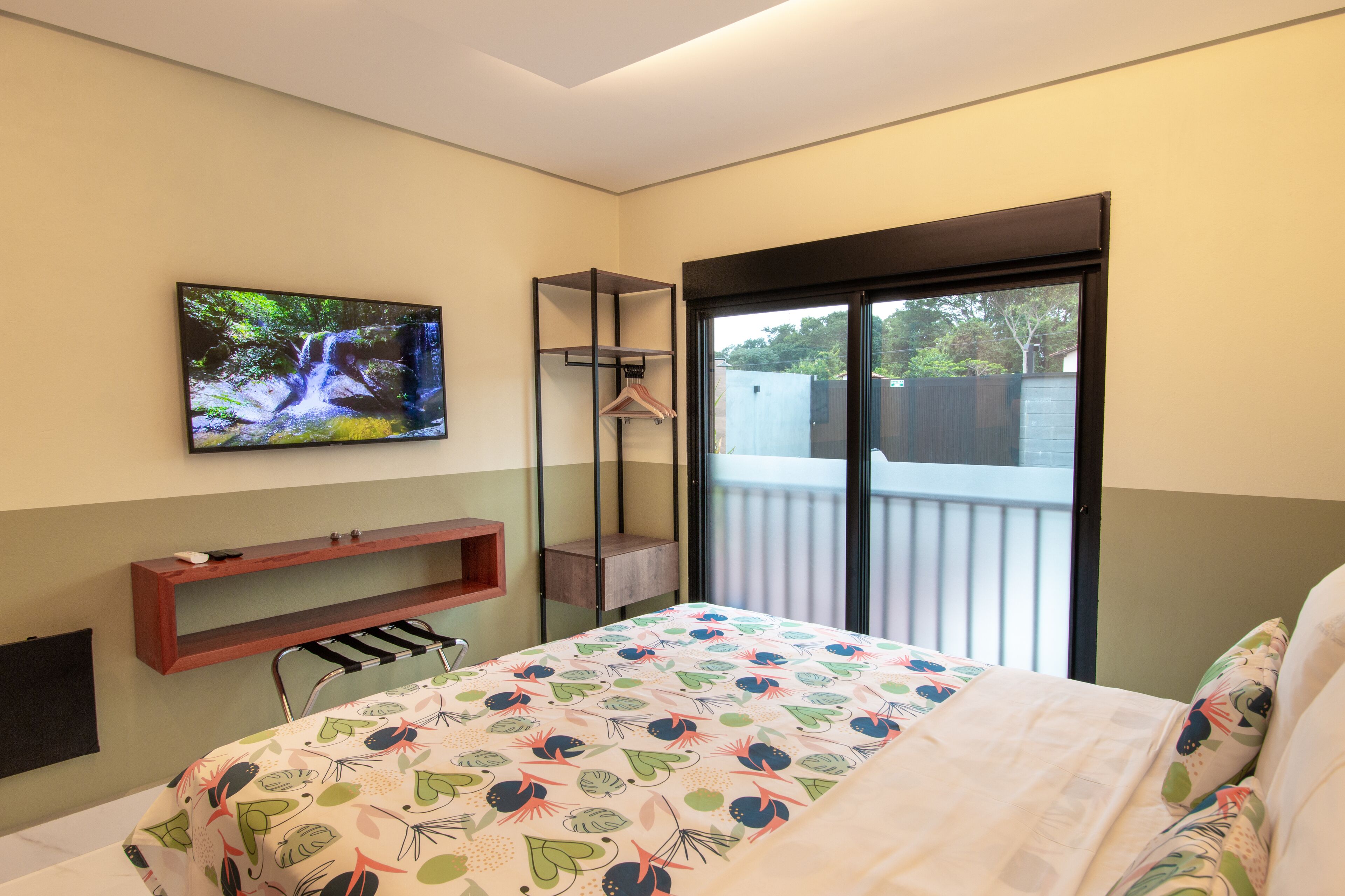 A bedroom with a bed, a TV mounted on the wall, a small wooden shelf, and a sliding glass door leading to a balcony.