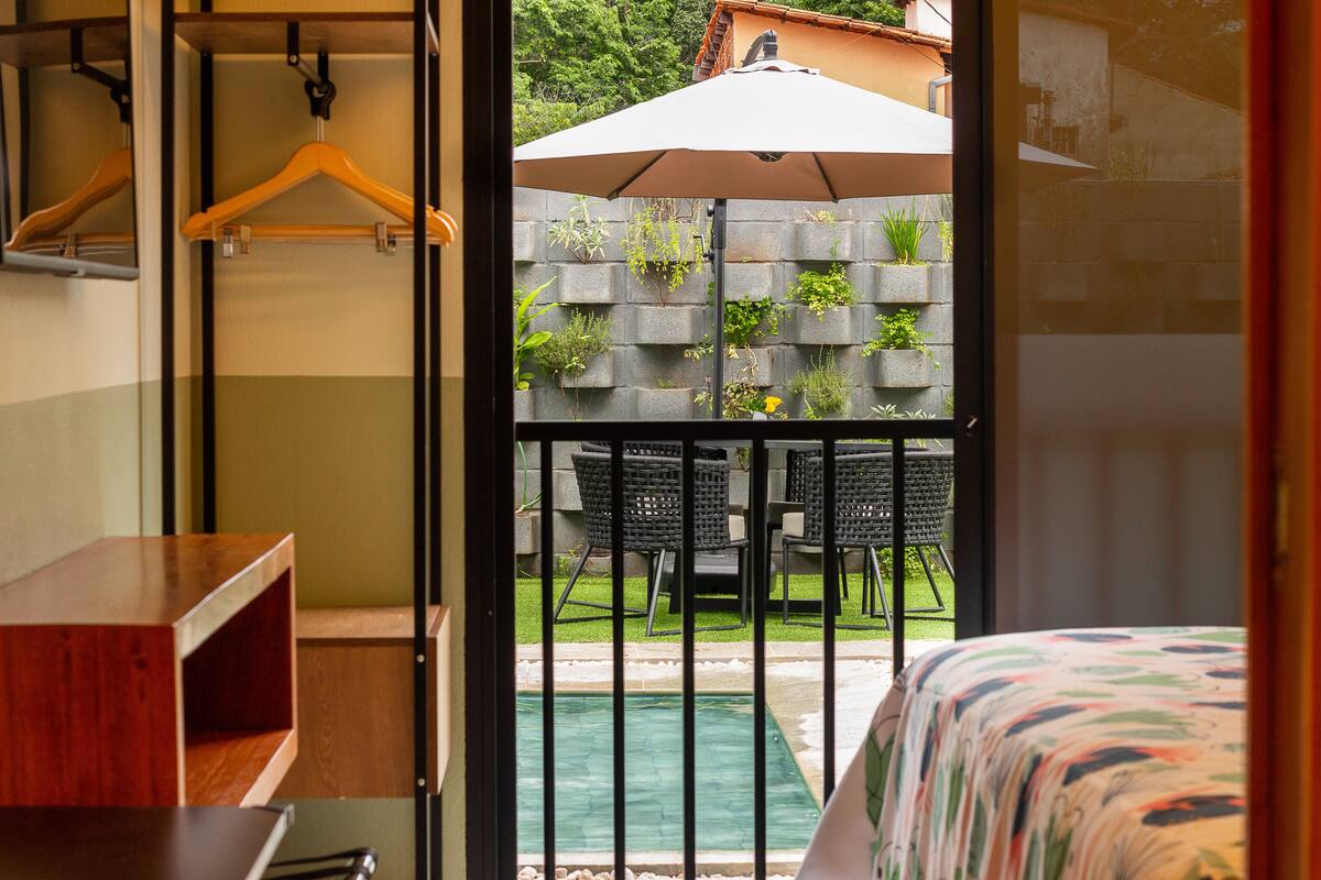 A room with a bed, a wooden dresser, a hanging rack, and a view of an outdoor area with a pool, chairs, and a hanging plant wall.