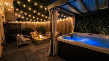 Private outdoor hot tub beneath a covered pergola accompanied by a fire pit, and BBQ grill underneath warm string lighting perfect for relaxing and unwinding in the evening.